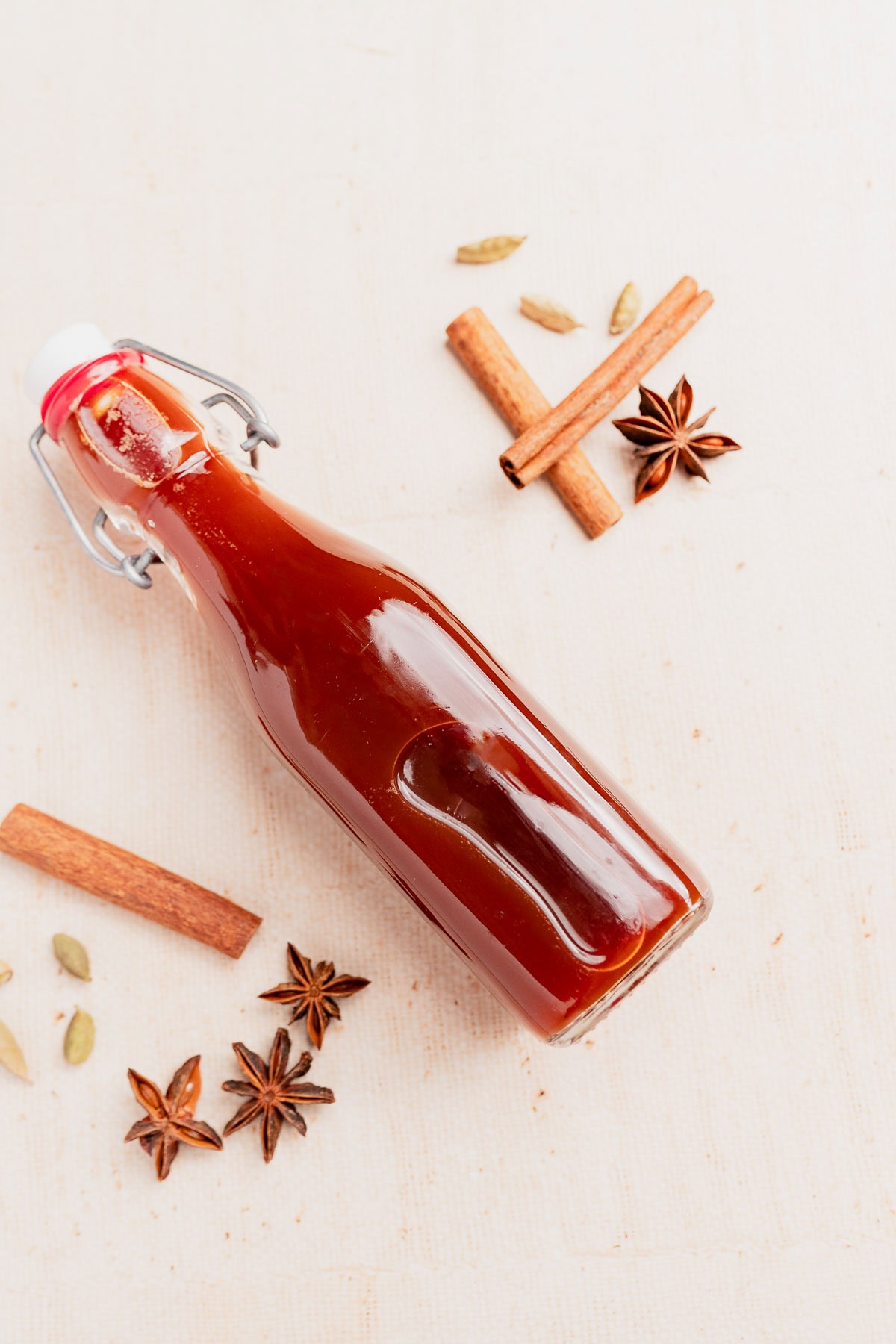 A glass bottle filled with brown chai concentrate, surrounded by cinnamon sticks, star anise, and cardamom pods on a light surface.