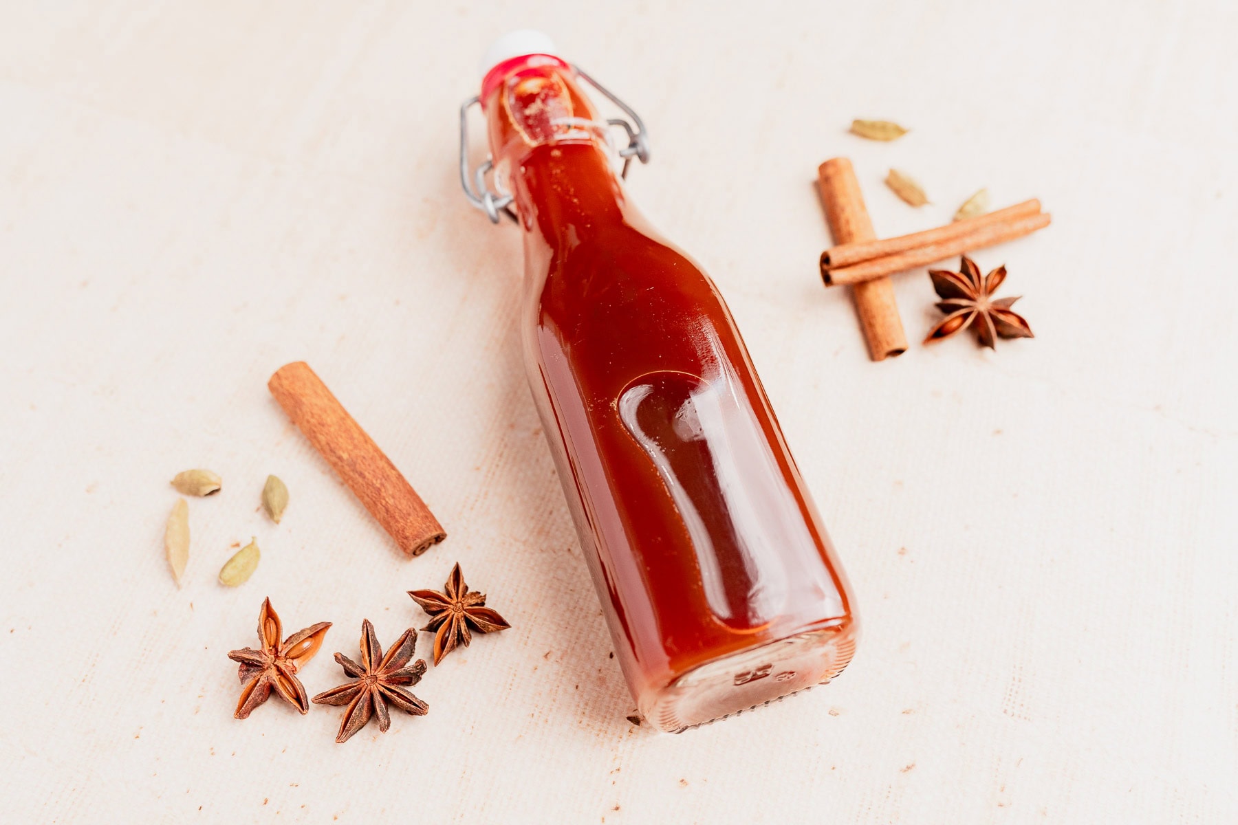 A glass bottle of chai concentrate with reddish-brown liquid lies on a light surface, surrounded by star anise, cinnamon sticks, and cardamom pods.