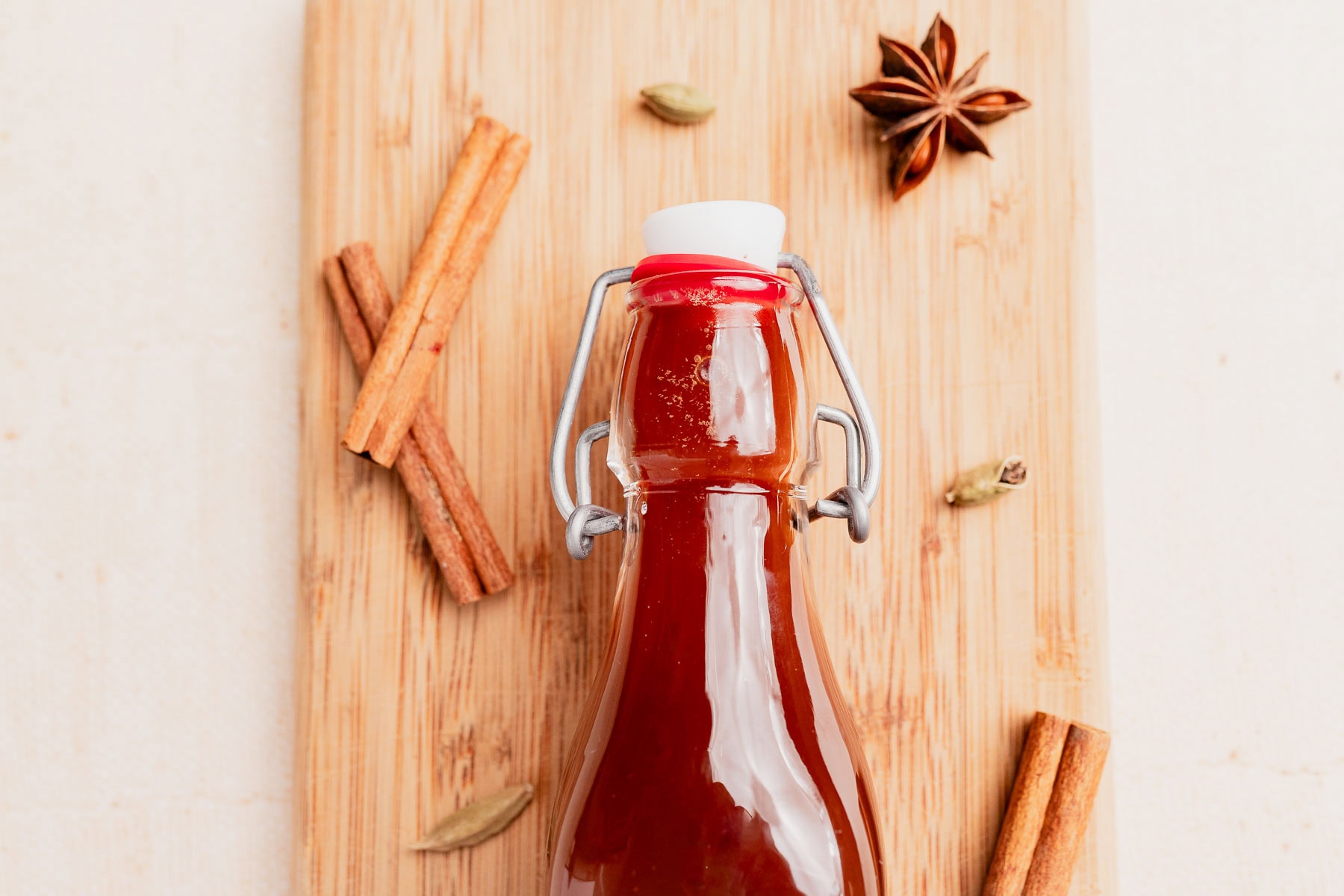A glass bottle of chai concentrate filled with reddish-brown liquid sits on a wooden board, surrounded by cinnamon sticks, star anise, and cardamom pods.