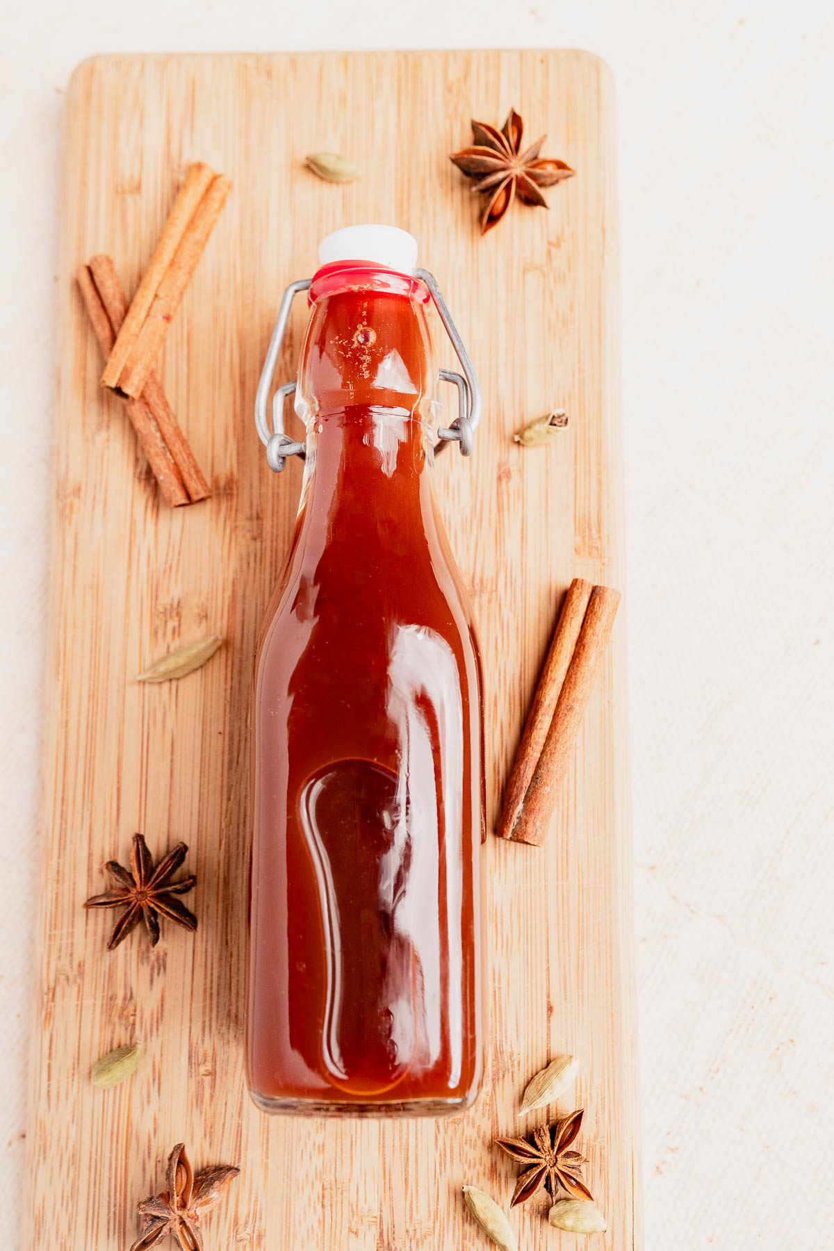 A glass bottle filled with reddish-brown chai concentrate sauce sits on a wooden board, surrounded by whole cinnamon sticks, star anise, and cardamom pods.