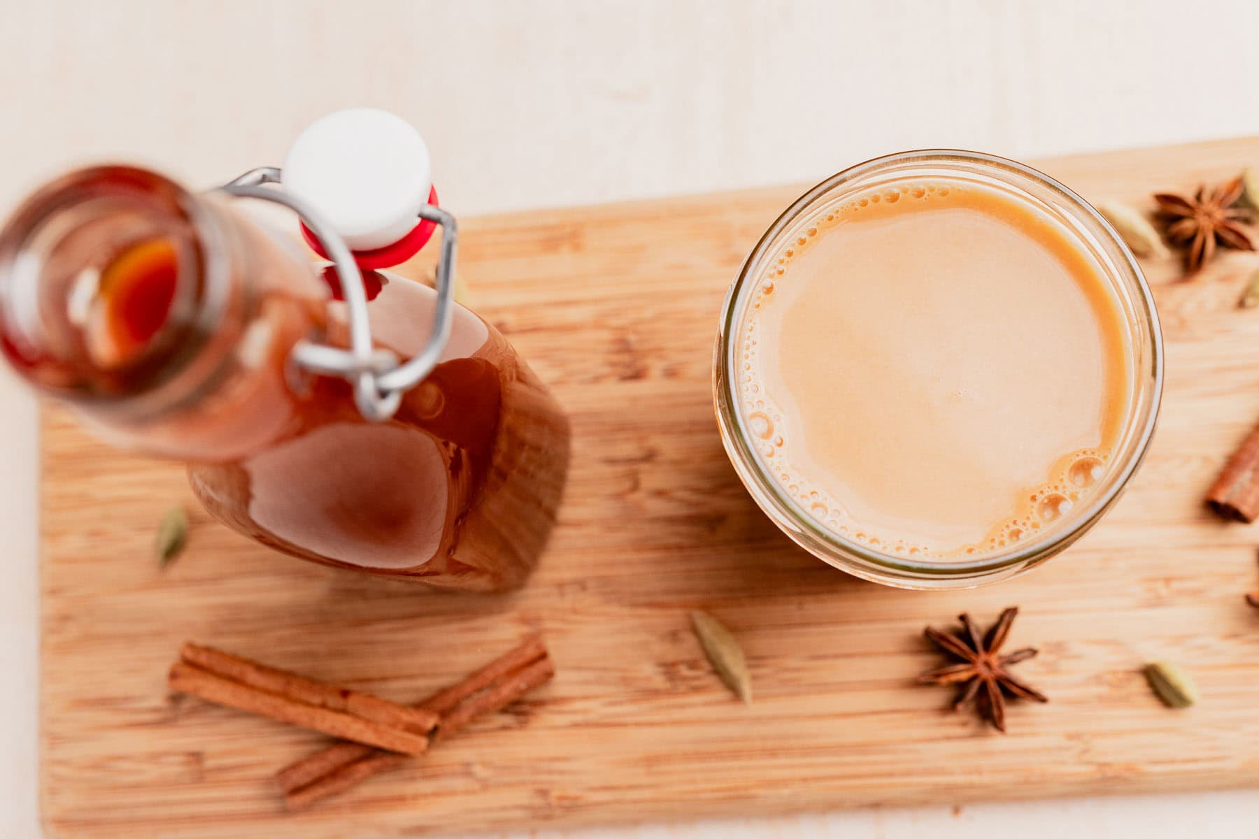 A glass and a bottle of chai concentrate sit on a wooden board, surrounded by cinnamon sticks, star anise, and cardamom pods, capturing the aromatic essence of chai concentrate in every detail.