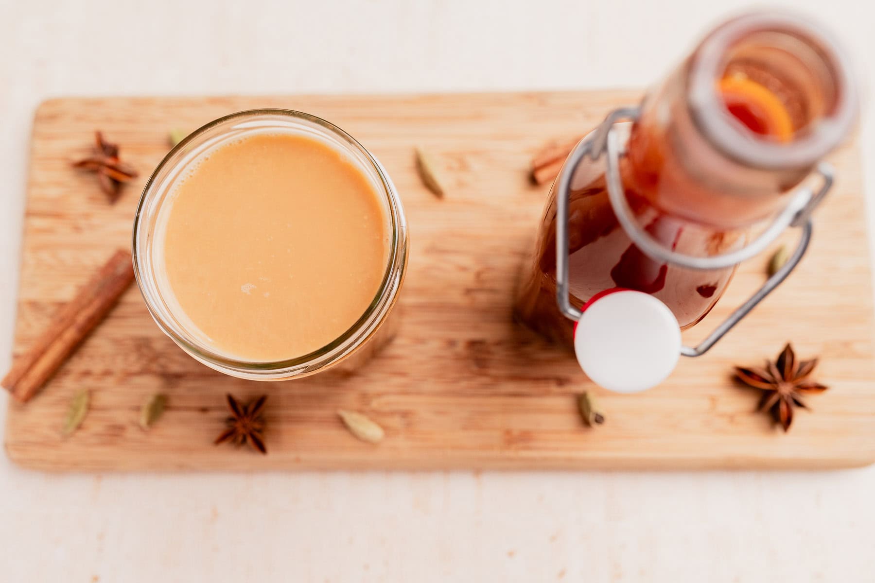 A glass of chai tea and a swing-top bottle of chai concentrate sit on a wooden board, surrounded by whole spices including star anise, cardamom pods, and a cinnamon stick.