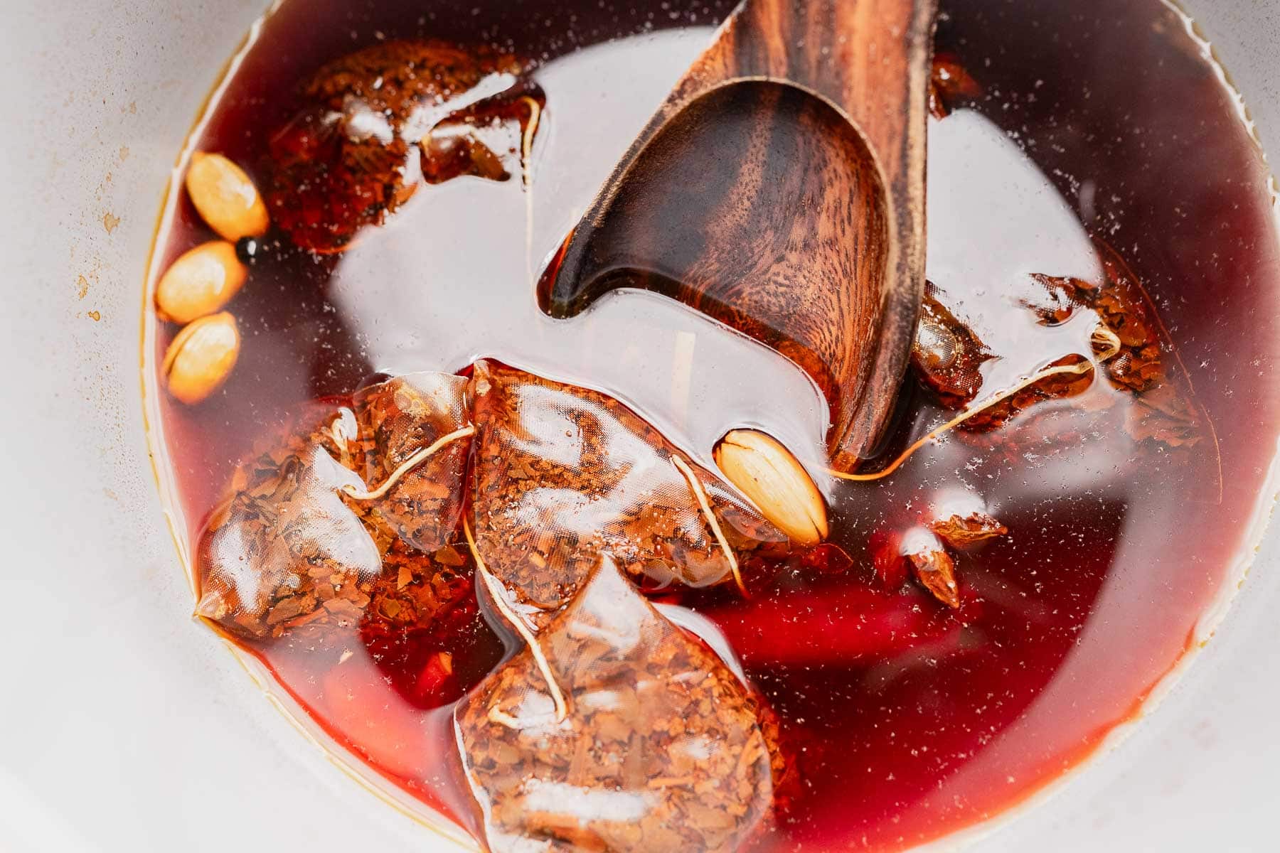 A wooden spoon stirs tea bags, spices, and whole nuts or seeds in a bowl of dark reddish chai concentrate.