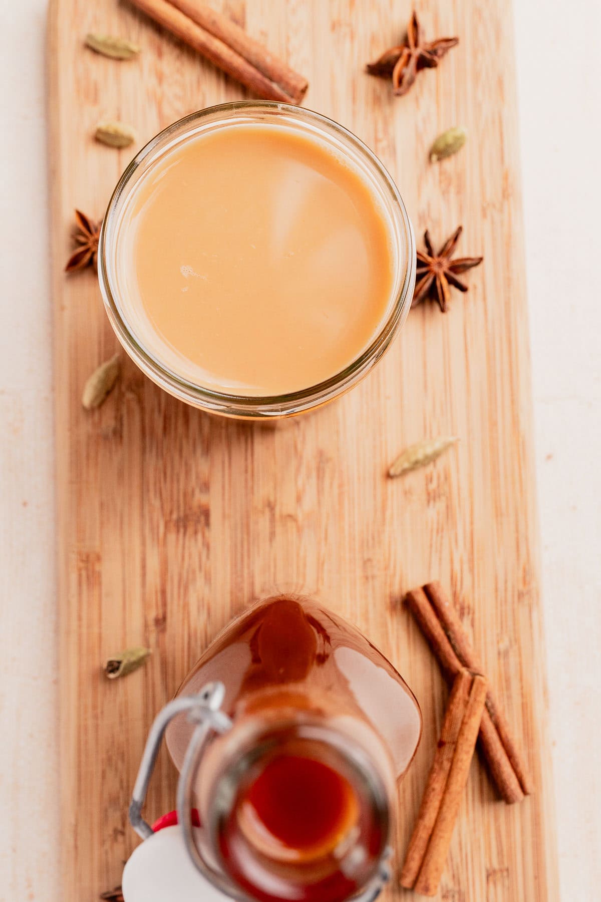 A glass of chai concentrate sits on a wooden board, surrounded by cinnamon sticks, star anise, and cardamom pods, with a bottle of rich chai concentrate placed nearby.