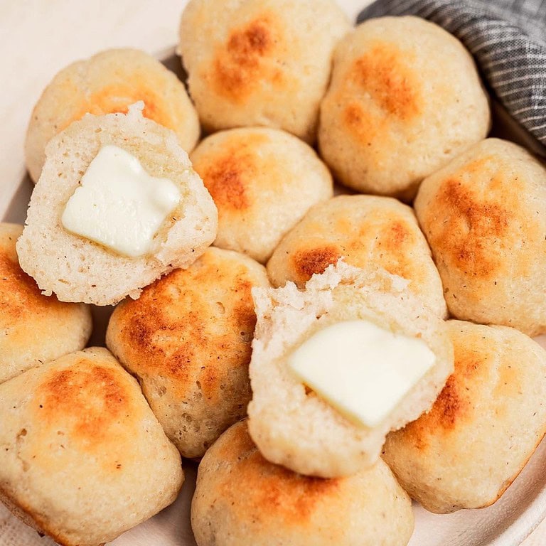 A plate of round, golden-brown gluten free dinner rolls, with two split open and topped with melting pats of butter. A gray striped cloth is partially visible in the corner.