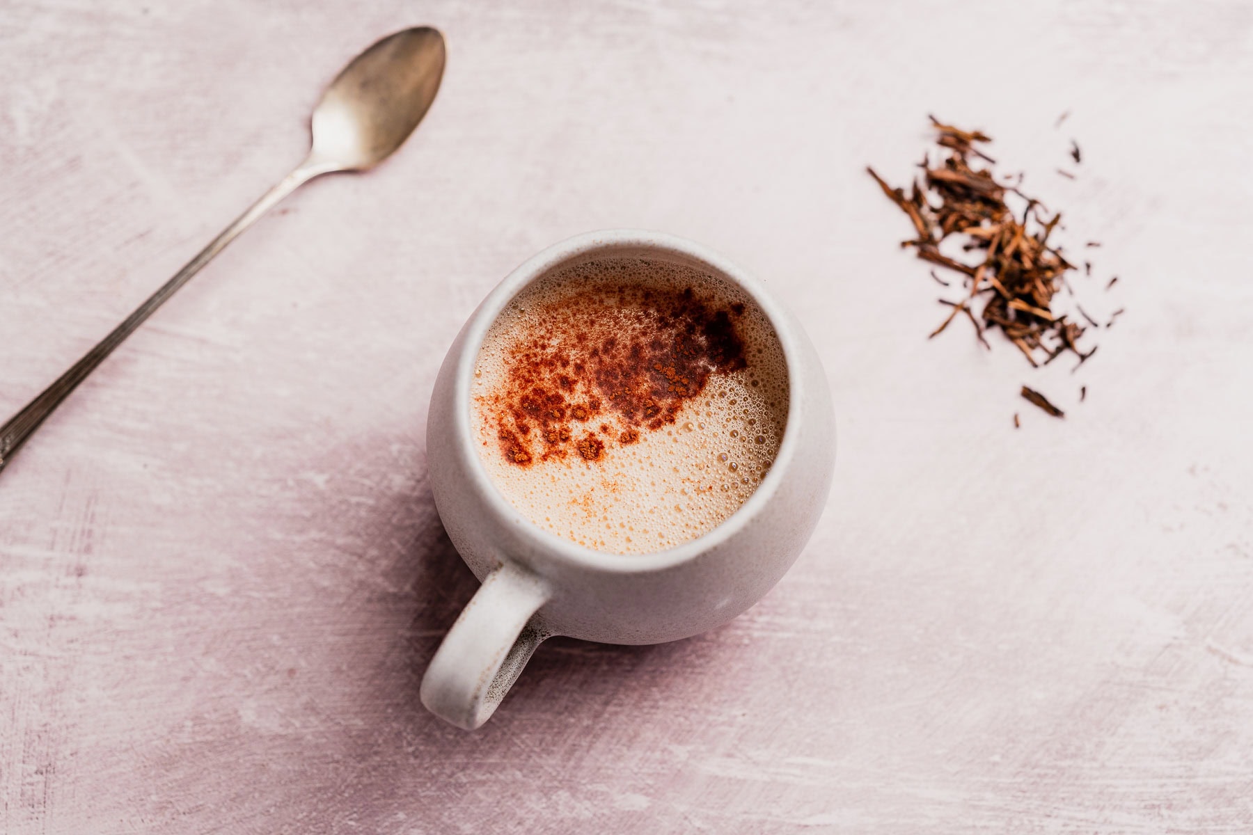 A white mug of frothy hojicha latte with a sprinkle of cinnamon sits beside a metal spoon and a small pile of loose tea leaves on a light pink surface.
