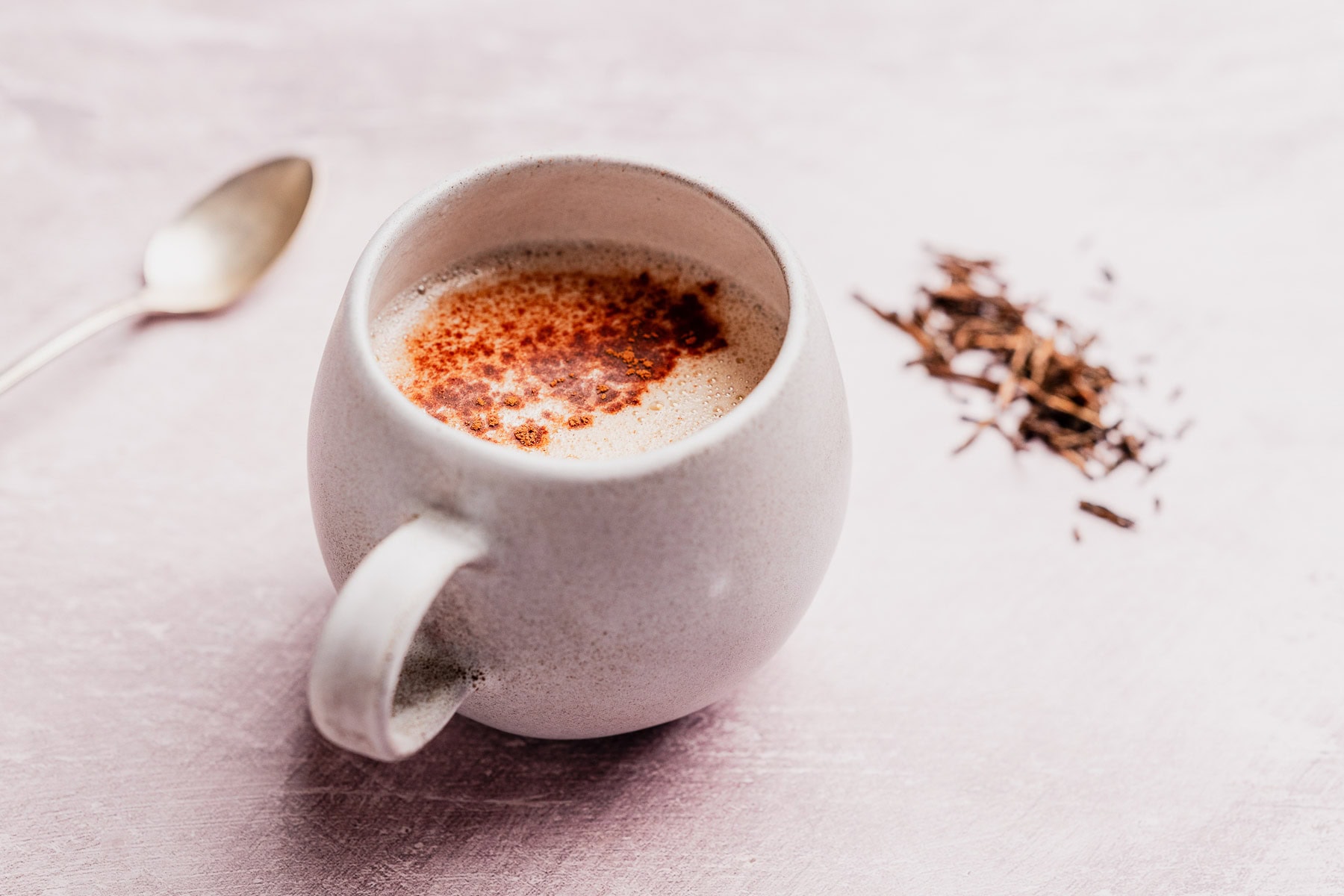 A white mug filled with chai tea topped with spices, placed beside a spoon and a small pile of loose hojicha latte tea leaves on a light surface.