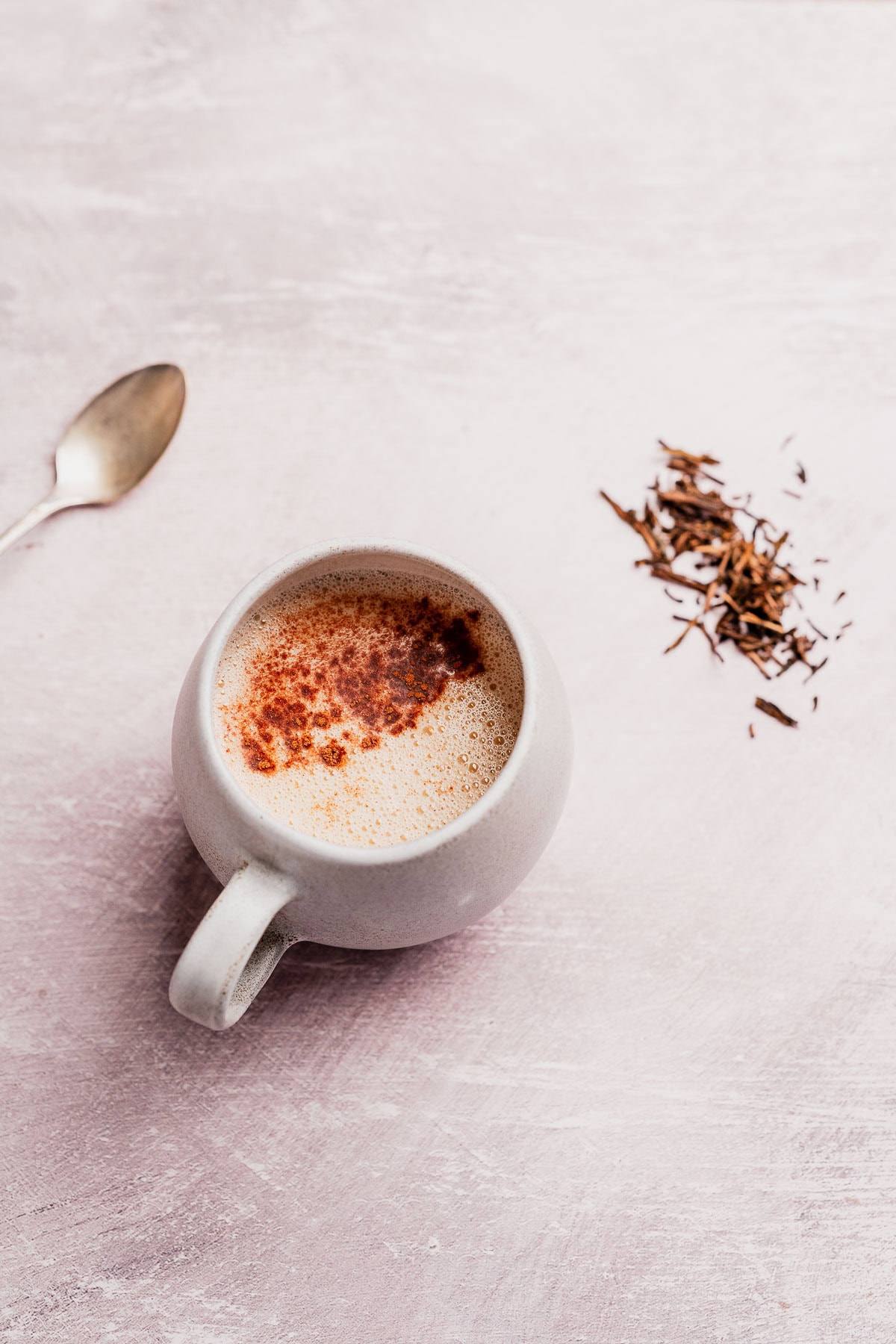 A cup of hojicha latte with a sprinkling of cinnamon, a spoon, and loose tea leaves on a light pink surface.