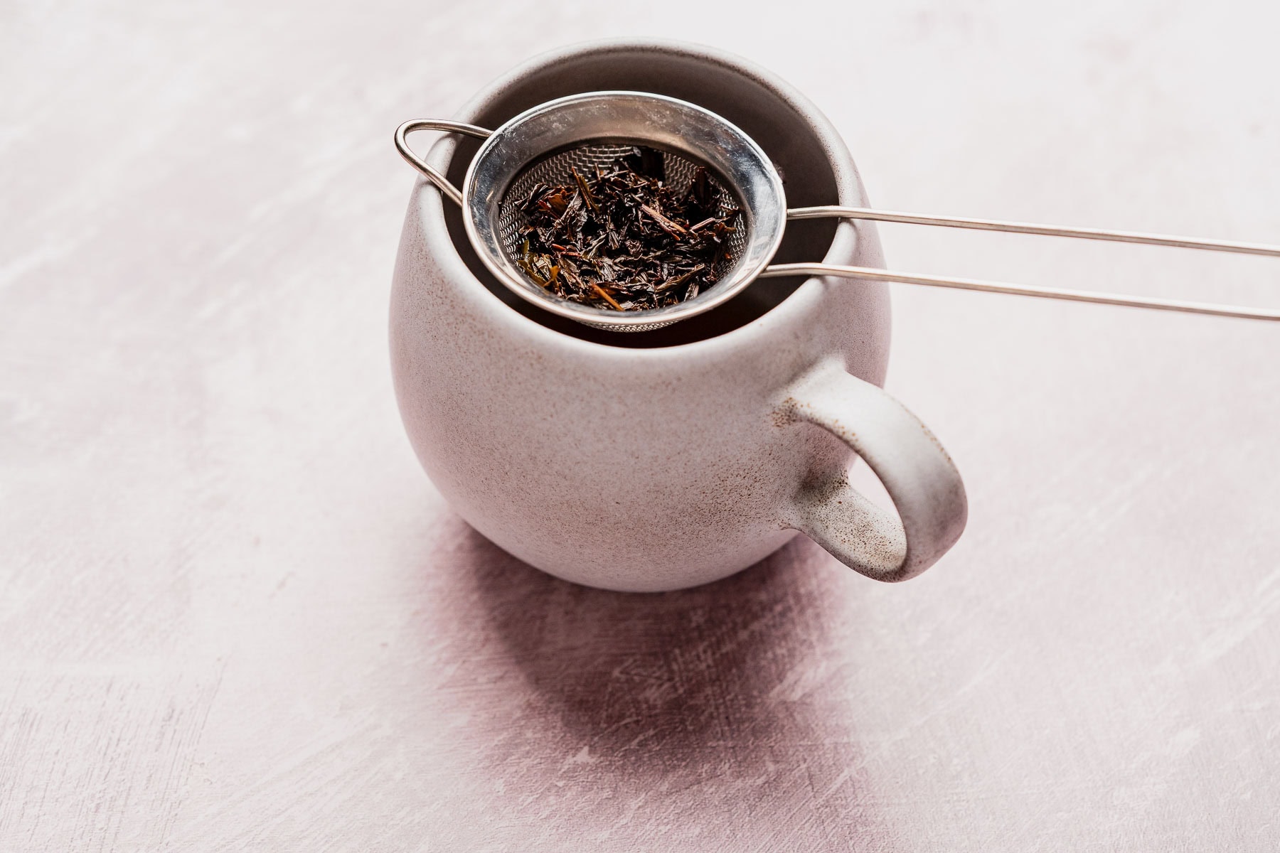 A white ceramic mug with a metal tea strainer holding loose hojicha latte tea leaves rests on top, set against a light, textured surface.