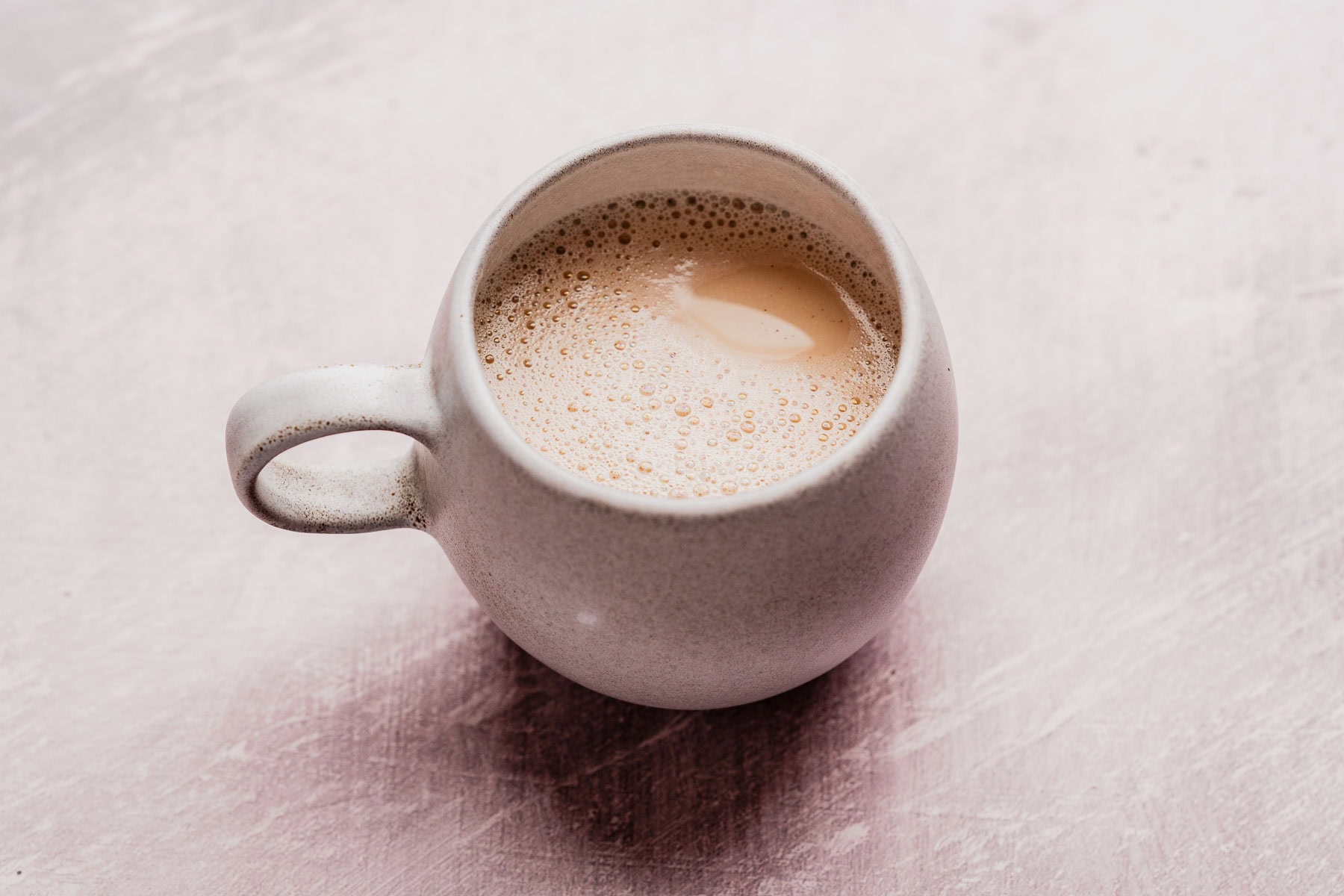 A white ceramic mug filled with a frothy hojicha latte sits on a light pink textured surface.