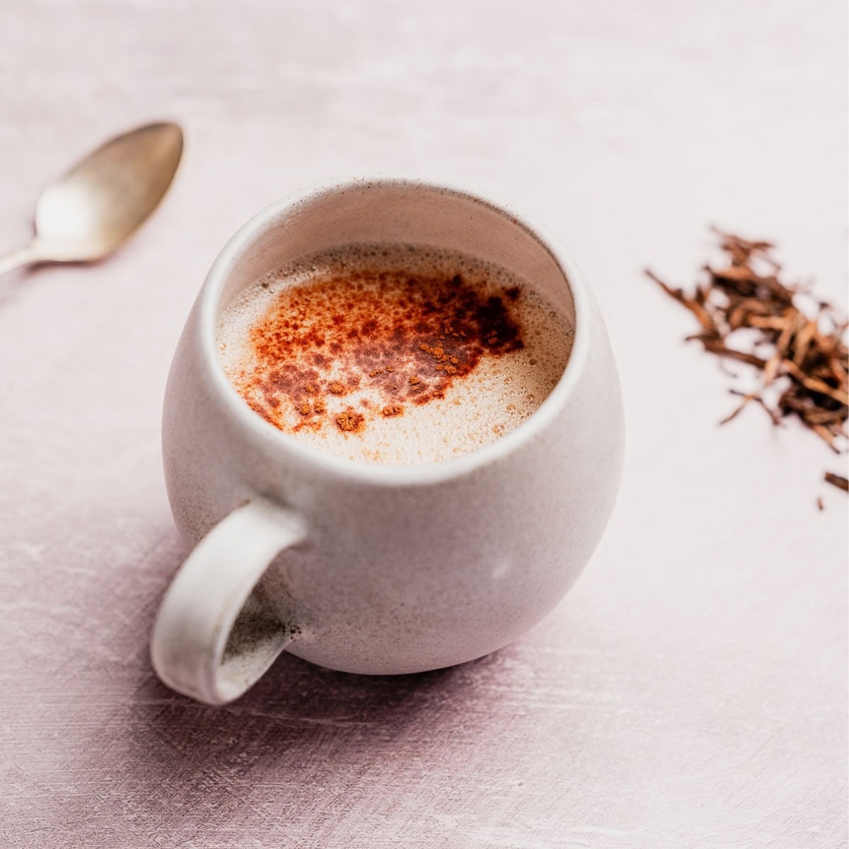 A white mug filled with a frothy hojicha latte topped with cinnamon sits on a light surface beside a spoon and a small pile of loose tea leaves.