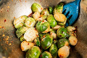 A metal bowl filled with roasted Brussels sprouts, some halved, mixed with red pepper flakes and oil, accompanied by a blue slotted spoon.