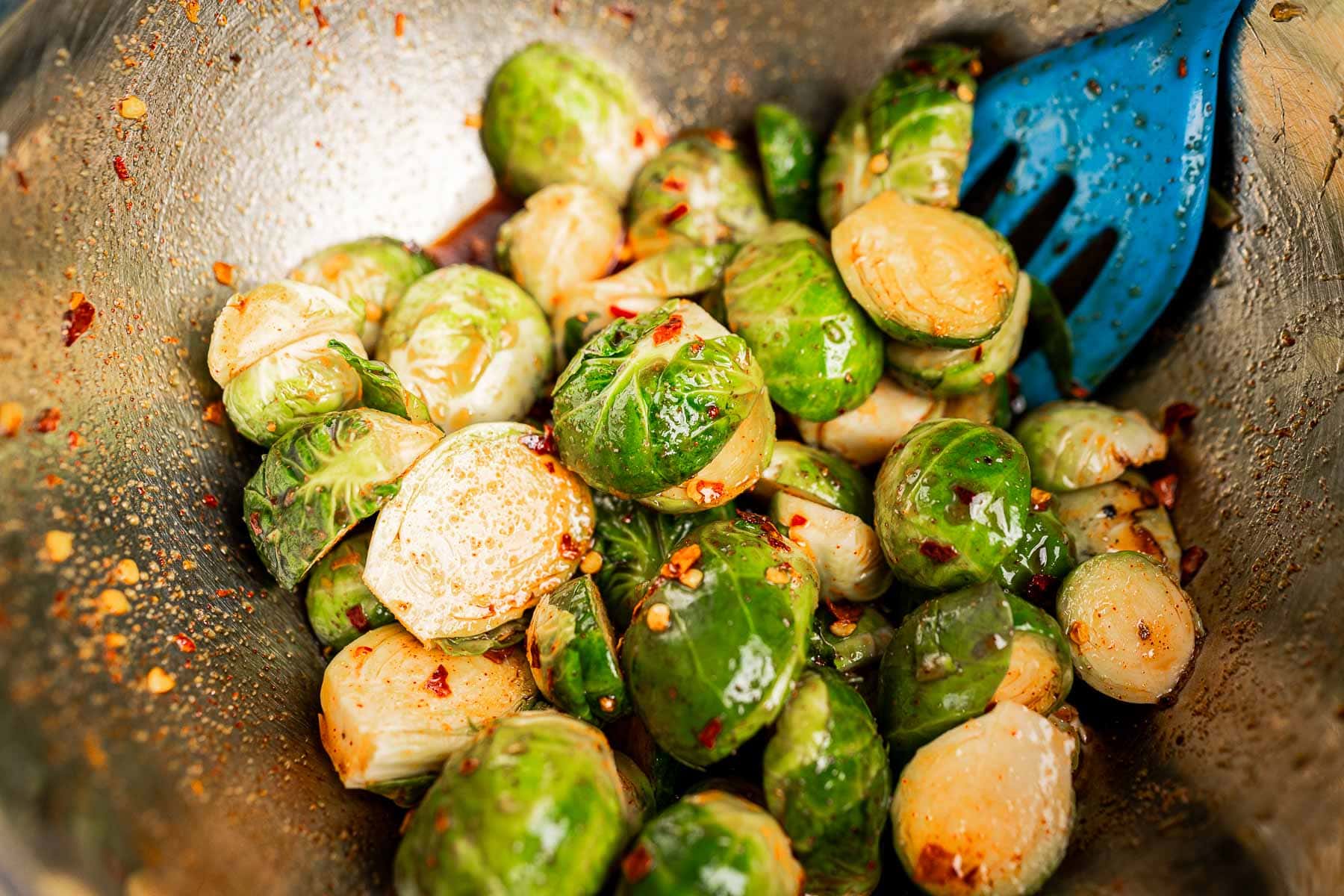 A metal bowl filled with roasted Brussels sprouts, some halved, mixed with red pepper flakes and oil, accompanied by a blue slotted spoon.