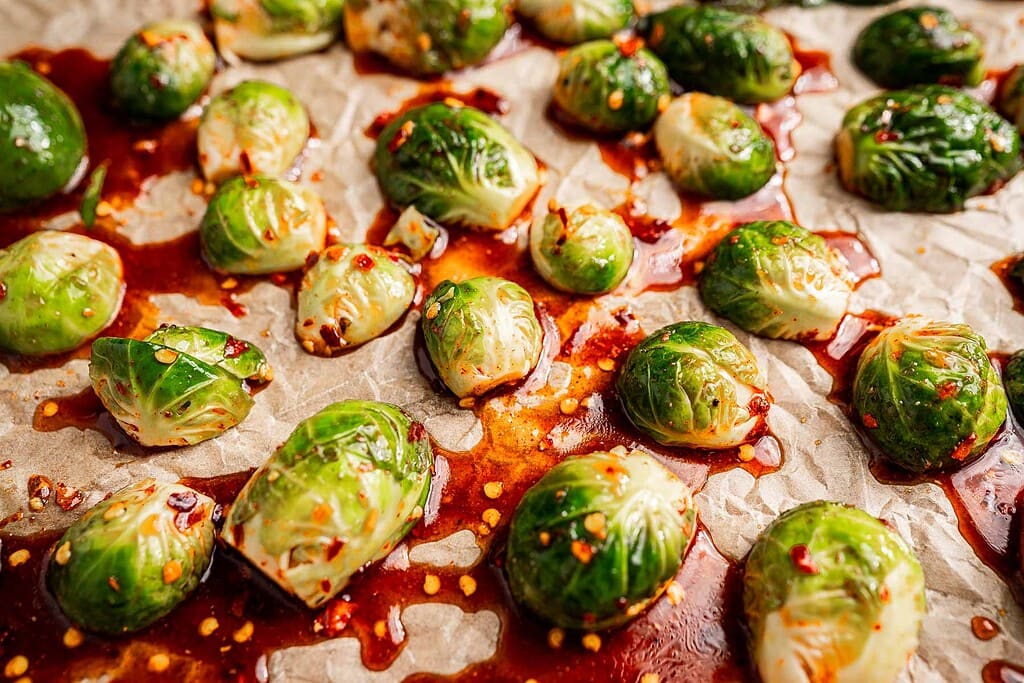 Halved Brussels sprouts on parchment paper, coated with a reddish sauce and sprinkled with chili flakes, ready to become delicious roasted brussels sprouts in the oven.