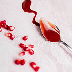 A spoon with red Pomegranate Salad Dressing rests on a light surface next to a wavy spill and scattered pomegranate seeds.