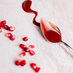 A spoon with red Pomegranate Salad Dressing rests on a light surface next to a wavy spill and scattered pomegranate seeds.