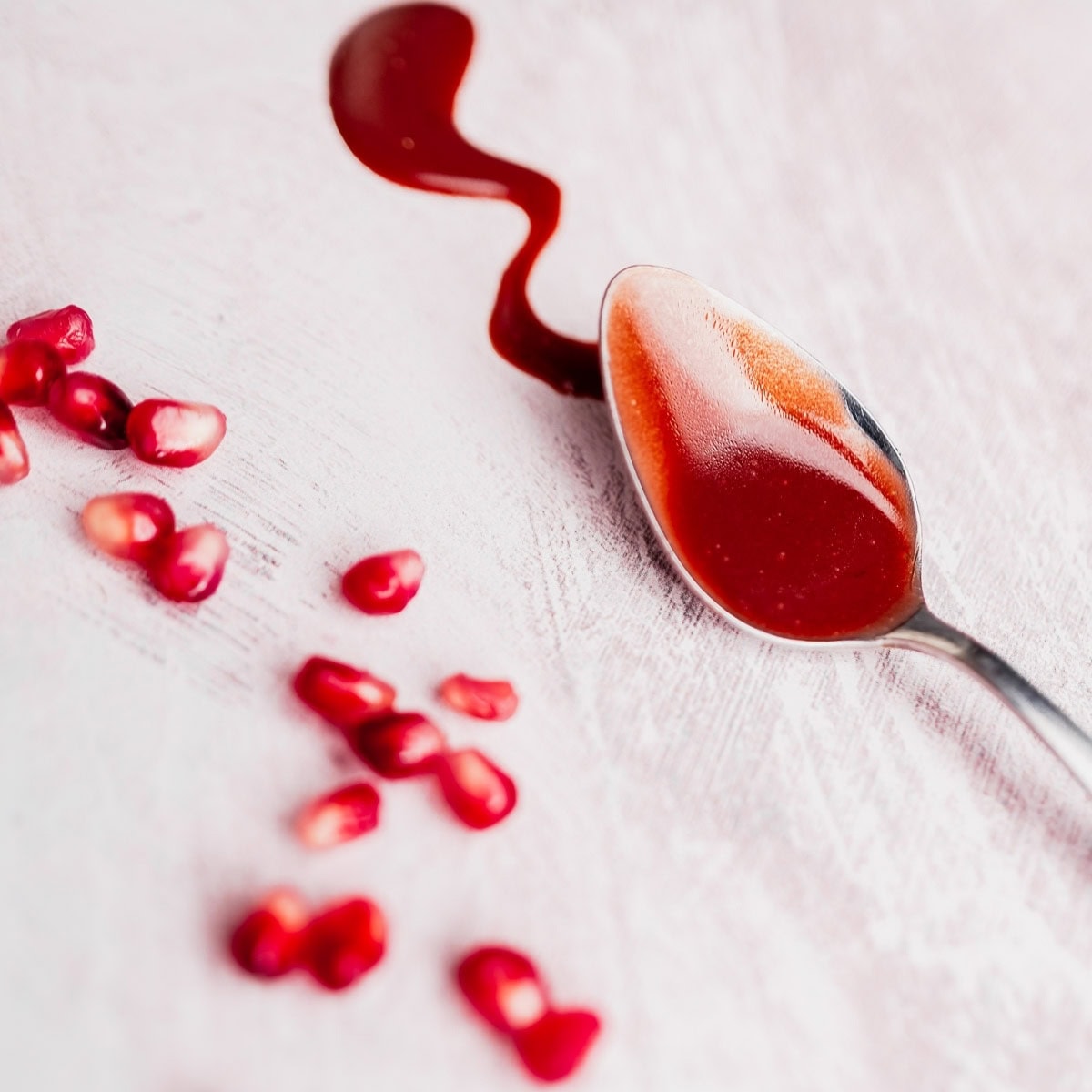 A spoon with red Pomegranate Salad Dressing rests on a light surface next to a wavy spill and scattered pomegranate seeds.