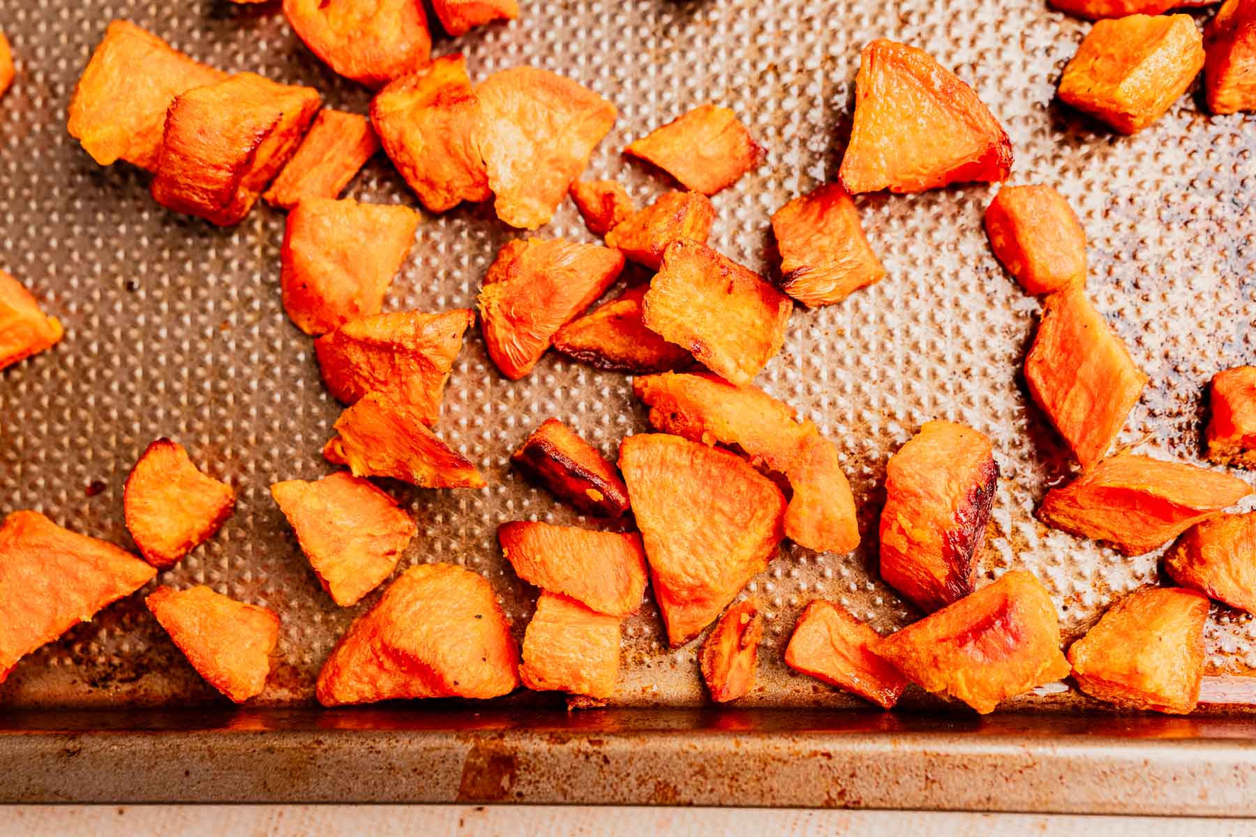 Chunks of roasted sweet potatoes, perfect for adding depth to pumpkin and sweet potato soup, scattered on a textured metal baking sheet.