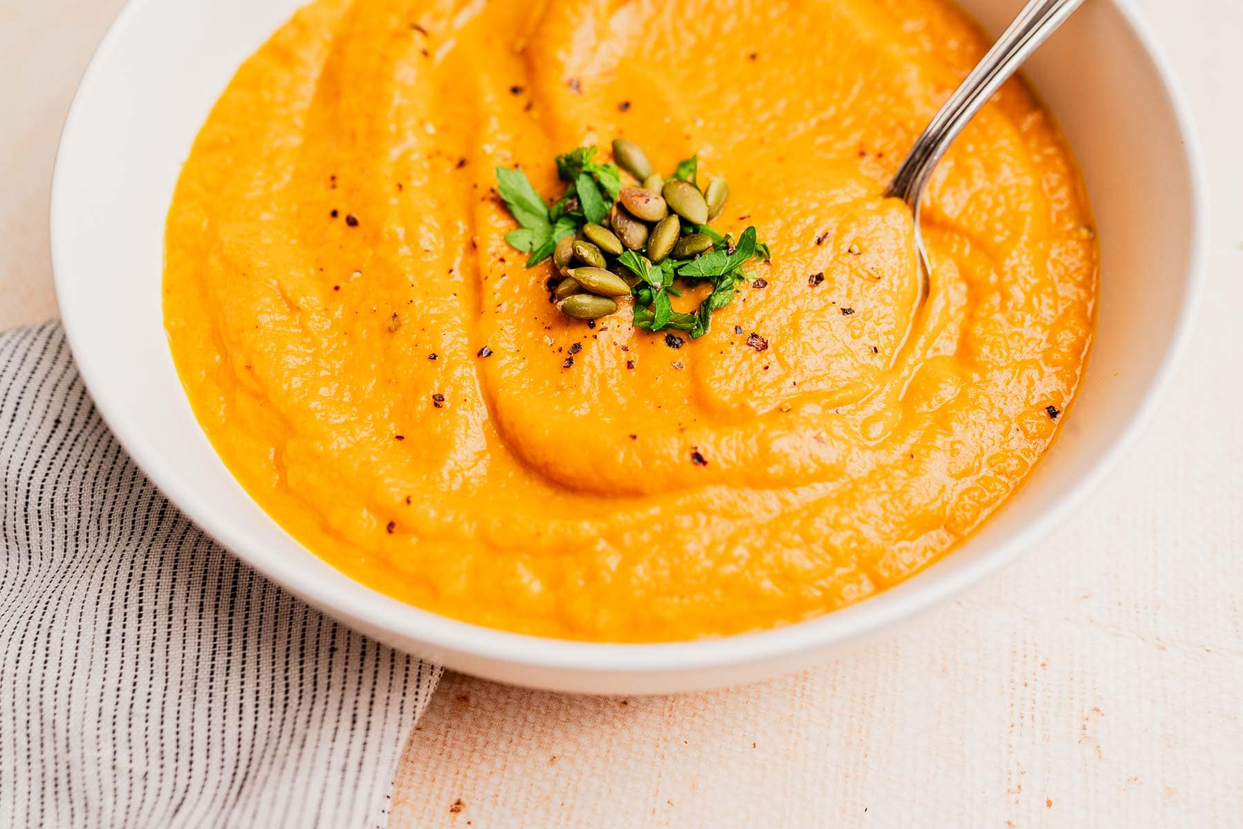 A bowl of pumpkin and sweet potato soup, orange in color, is garnished with chopped herbs and pumpkin seeds, with a spoon resting in the bowl.