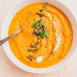 A bowl of pumpkin and sweet potato soup garnished with pumpkin seeds, fresh parsley, black pepper, and a swirl of cream, with a spoon resting in the bowl.