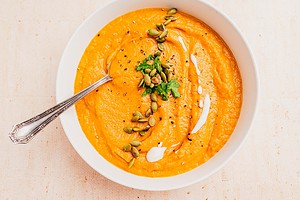 A bowl of pumpkin and sweet potato soup garnished with pumpkin seeds, fresh parsley, black pepper, and a swirl of cream, with a spoon resting in the bowl.