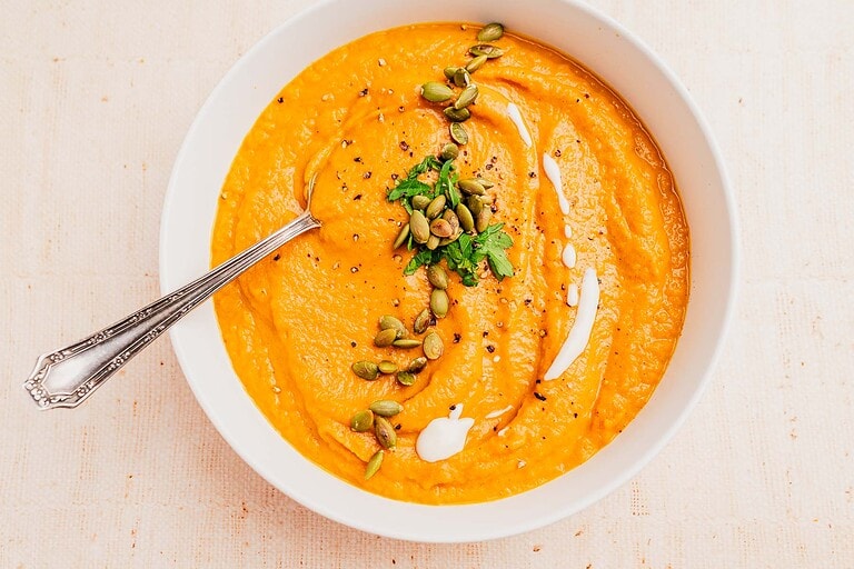 A bowl of pumpkin and sweet potato soup garnished with pumpkin seeds, fresh parsley, black pepper, and a swirl of cream, with a spoon resting in the bowl.