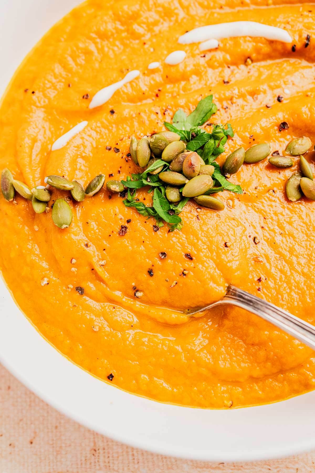 A bowl of creamy pumpkin and sweet potato soup garnished with pumpkin seeds, chopped herbs, ground pepper, and a swirl of cream, with a spoon resting in the bowl.