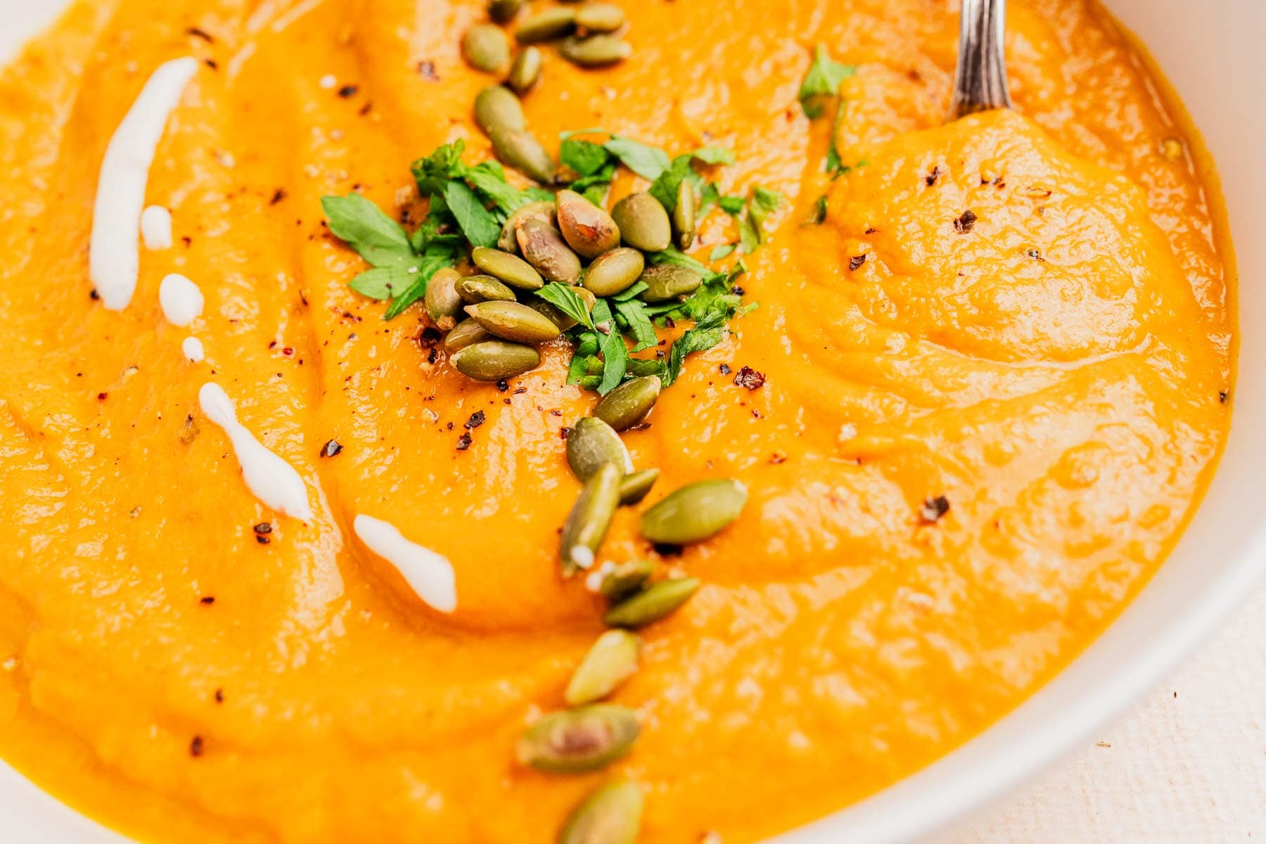 A bowl of creamy pumpkin and sweet potato soup garnished with pumpkin seeds, chopped herbs, ground pepper, and a drizzle of cream, with a spoon partially visible.