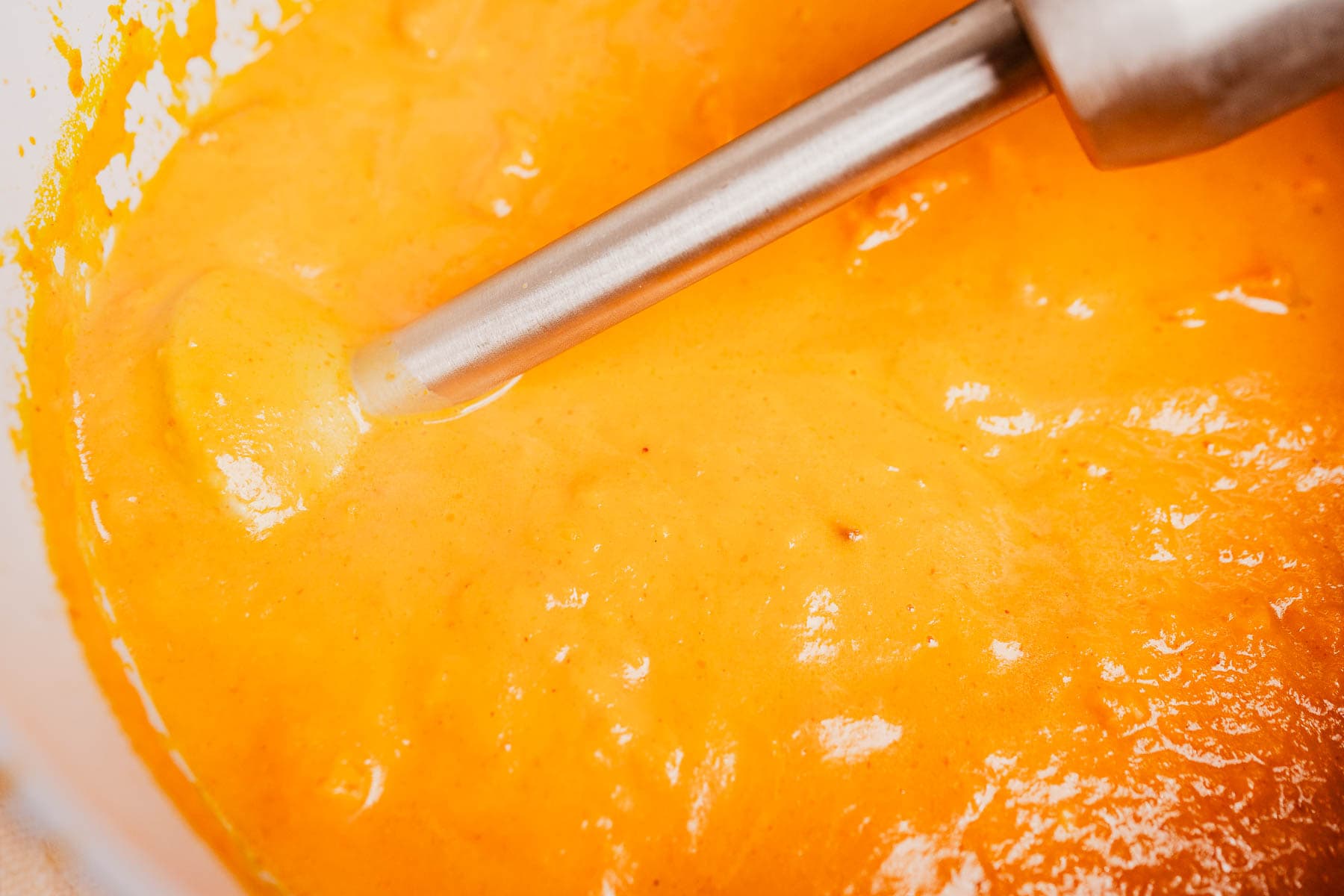A close-up of an immersion blender blending a smooth, orange pumpkin and sweet potato soup mixture in a bowl.