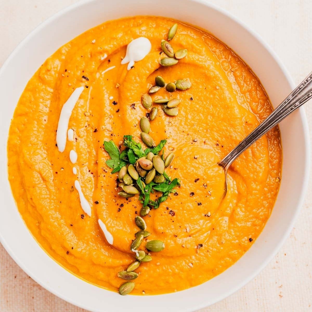A bowl of creamy pumpkin and sweet potato soup topped with pumpkin seeds, chopped herbs, a drizzle of cream, and black pepper, with a spoon inside.