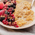 A bowl of quinoa breakfast porridge topped with blackberries, raspberries, strawberries, blueberries, and shredded coconut, served with a spoon on the side.