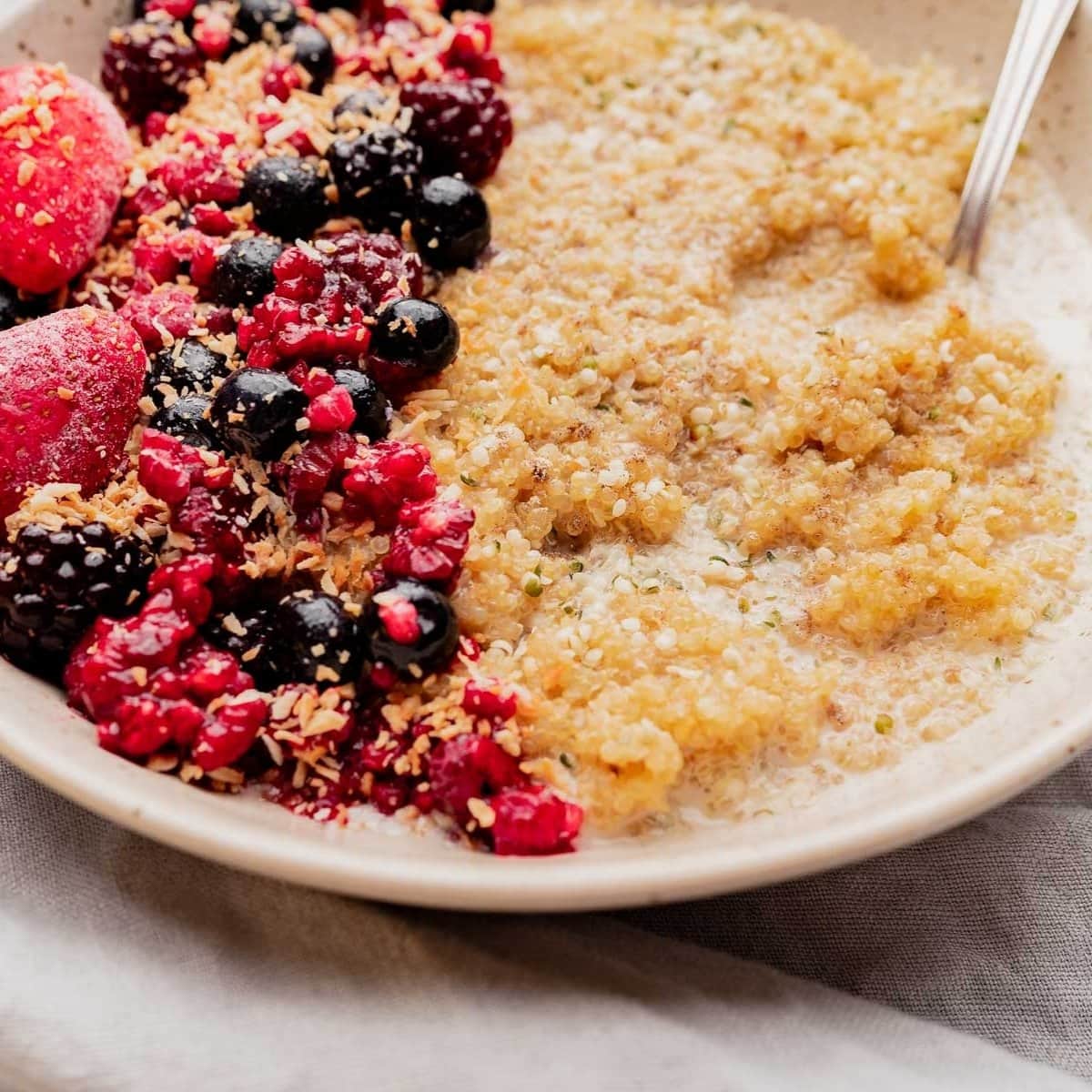 A bowl of quinoa breakfast porridge topped with blackberries, raspberries, strawberries, blueberries, and shredded coconut, served with a spoon on the side.