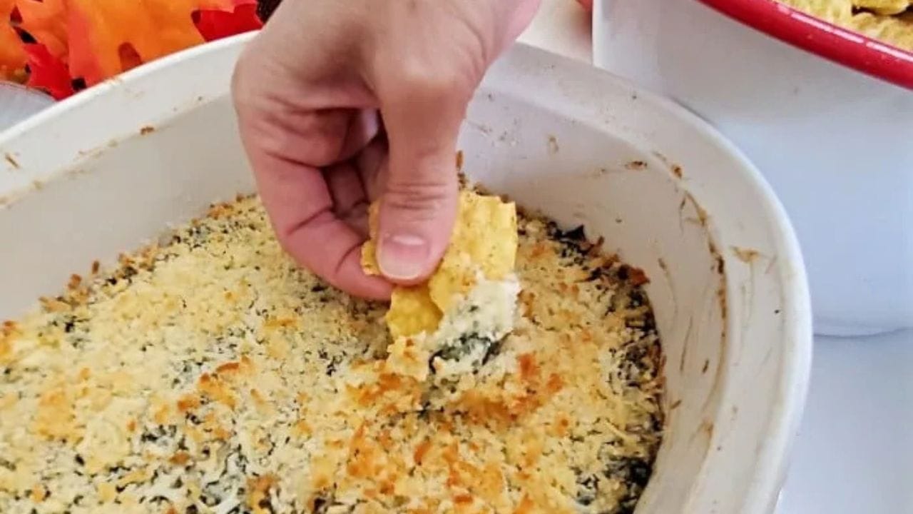 This photo shows a hand scooping out baking spinach artichoke dip from a white casserole dish.