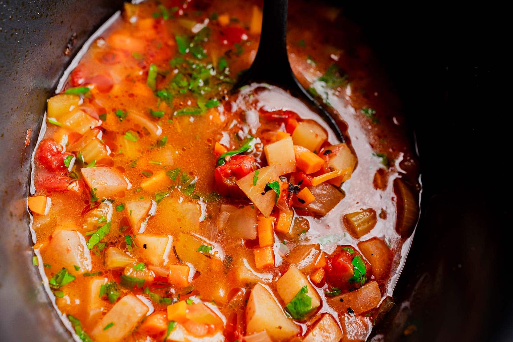 A ladle in a pot of slow cooker vegetable stew brimming with diced carrots, potatoes, tomatoes, and fresh herbs in a savory broth.