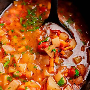 A close-up of a ladle serving chunky slow cooker vegetable stew with carrots, potatoes, tomatoes, and herbs in a flavorful broth.