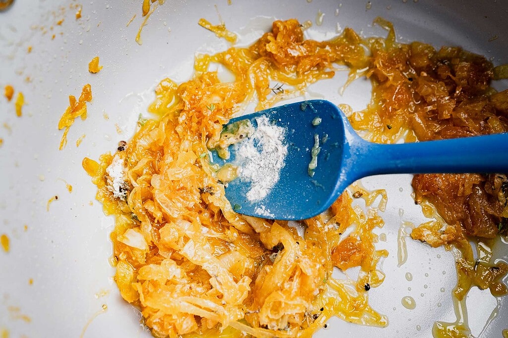 A blue spatula mixing cooked onions and powder in a white pan with some oil, the perfect base for vegetarian French onion soup.