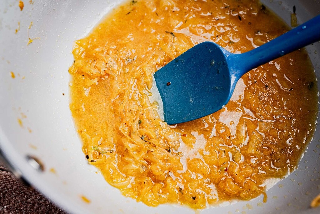 A blue spatula stirs caramelized onions in a white pan, creating a golden, saucy mixture perfect as the base for vegetarian French onion soup.
