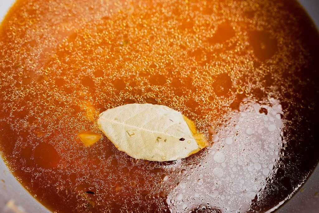 A close-up of reddish-brown broth with oil droplets and a single bay leaf floating on the surface, showcasing a warm bowl of vegetarian French onion soup.