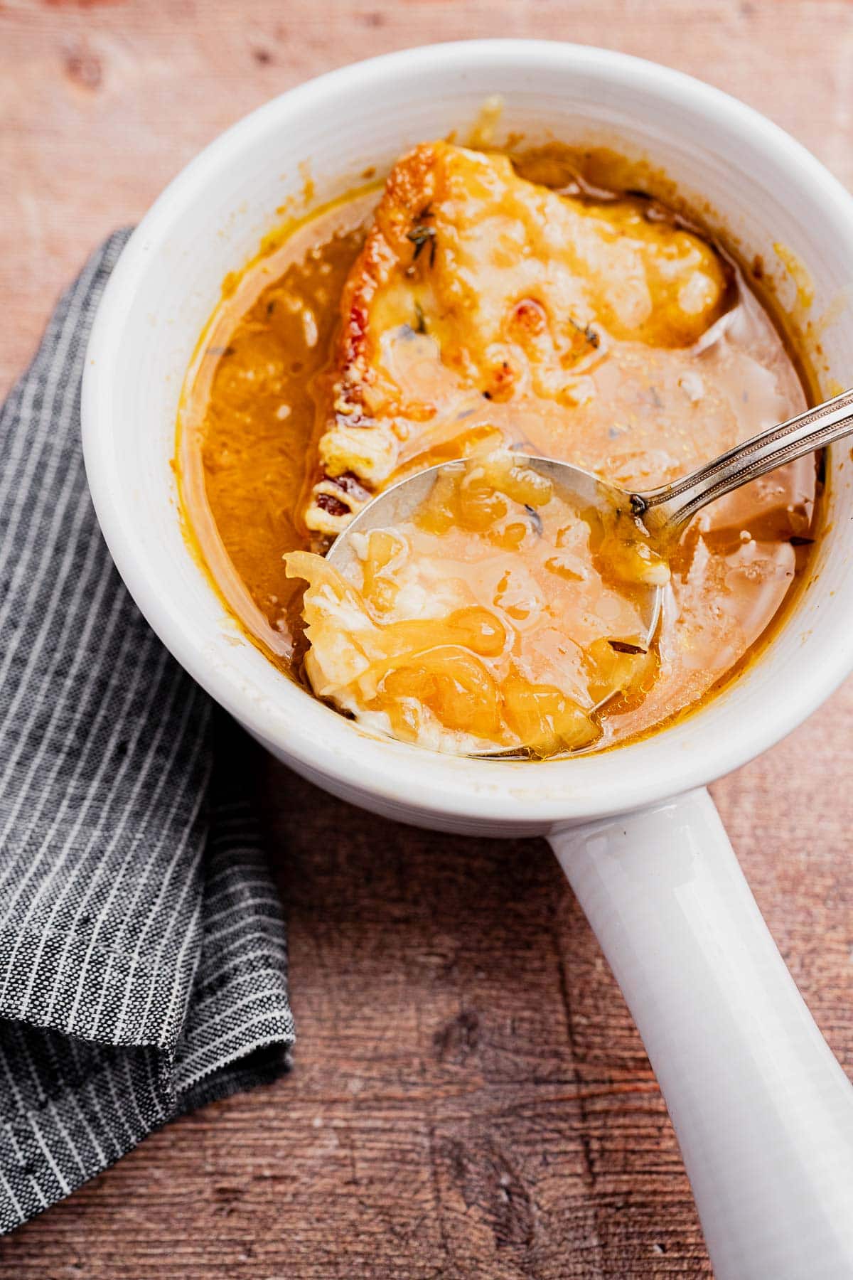 A bowl of vegetarian French onion soup topped with melted cheese and toasted bread sits on a wooden surface, with a spoon resting inside. A folded striped napkin is beside the bowl.