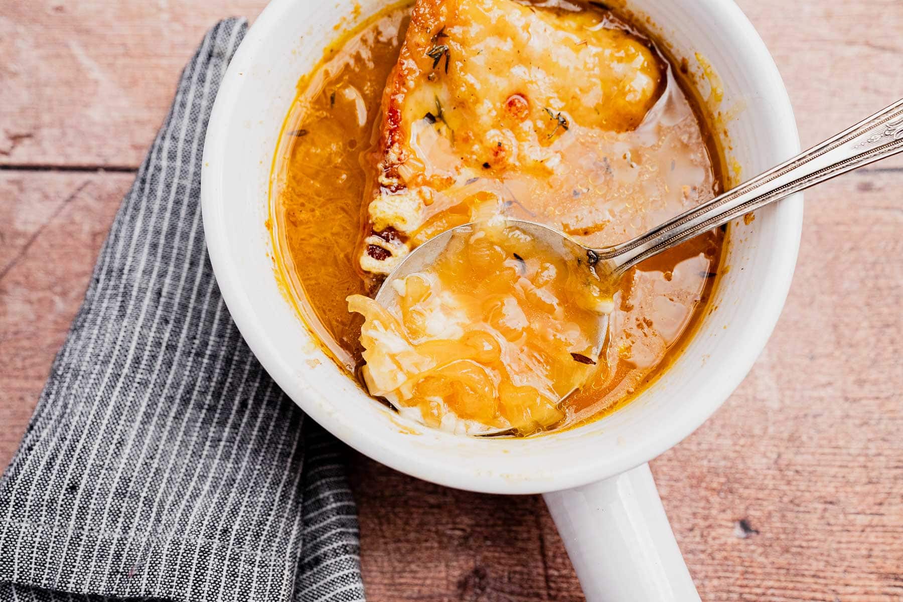 A bowl of vegetarian French onion soup topped with melted cheese and bread, with a spoon resting inside and a striped cloth napkin beside the bowl.