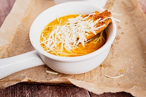 White bowl of vegetarian French onion soup with shredded cheese, thin noodles, and a slice of toasted bread on parchment paper.