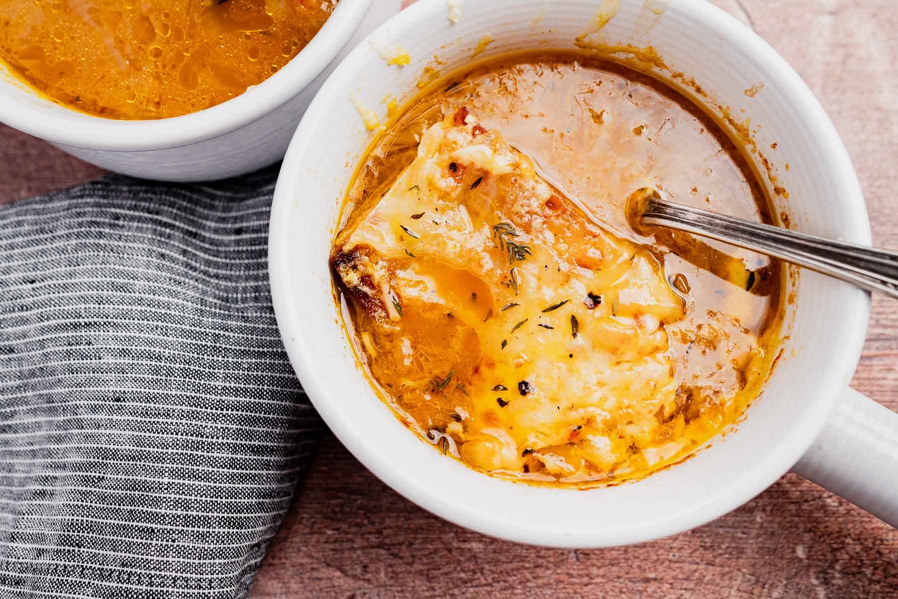A white bowl of vegetarian French onion soup topped with melted cheese and a slice of bread, with a spoon resting inside; another bowl and a striped napkin are nearby.