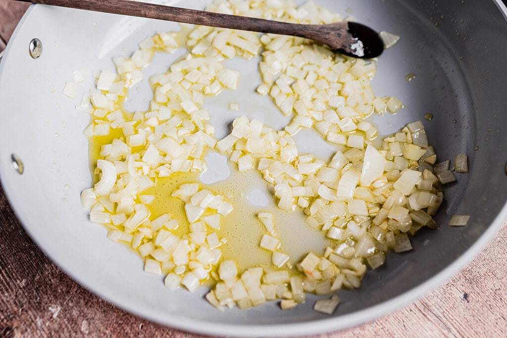 Chopped onions are sautéing in oil in a skillet for a hearty vegetarian shepherd's pie, with a wooden spoon resting on the edge of the pan.
