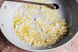 Chopped onions are sautéing in oil in a skillet for a hearty vegetarian shepherd's pie, with a wooden spoon resting on the edge of the pan.