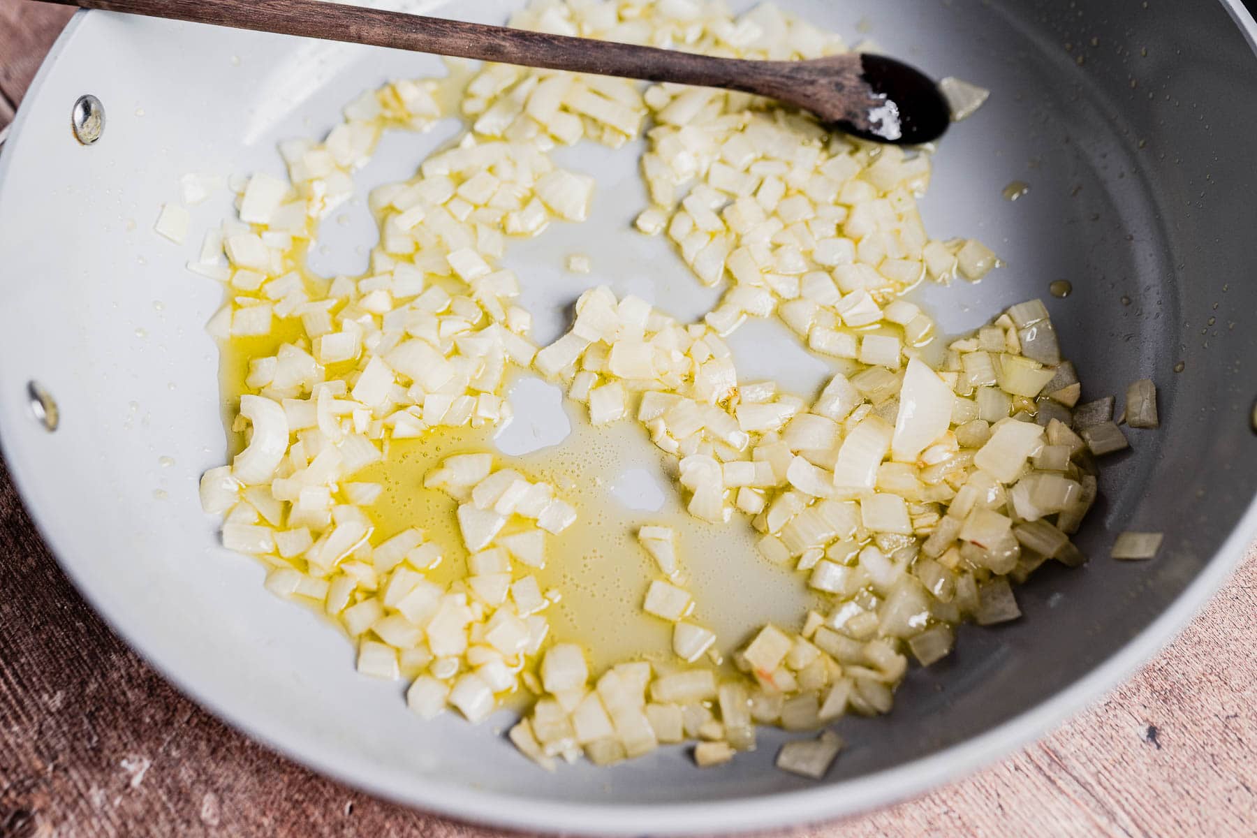 Chopped onions are sautéing in oil in a skillet for a hearty vegetarian shepherd's pie, with a wooden spoon resting on the edge of the pan.