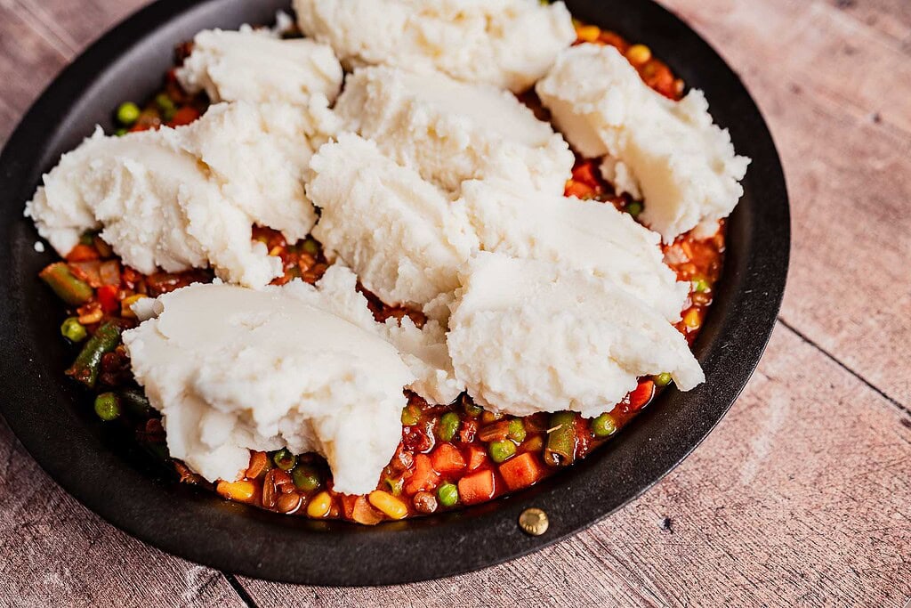 A round baking dish filled with mixed vegetables in sauce, topped with scoops of mashed potatoes, ready to be spread and baked into a comforting vegetarian shepherd's pie.