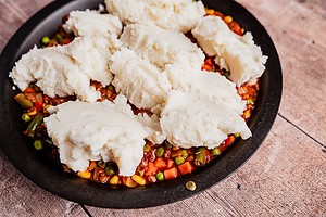A round baking dish filled with mixed vegetables in sauce, topped with scoops of mashed potatoes, ready to be spread and baked into a comforting vegetarian shepherd's pie.