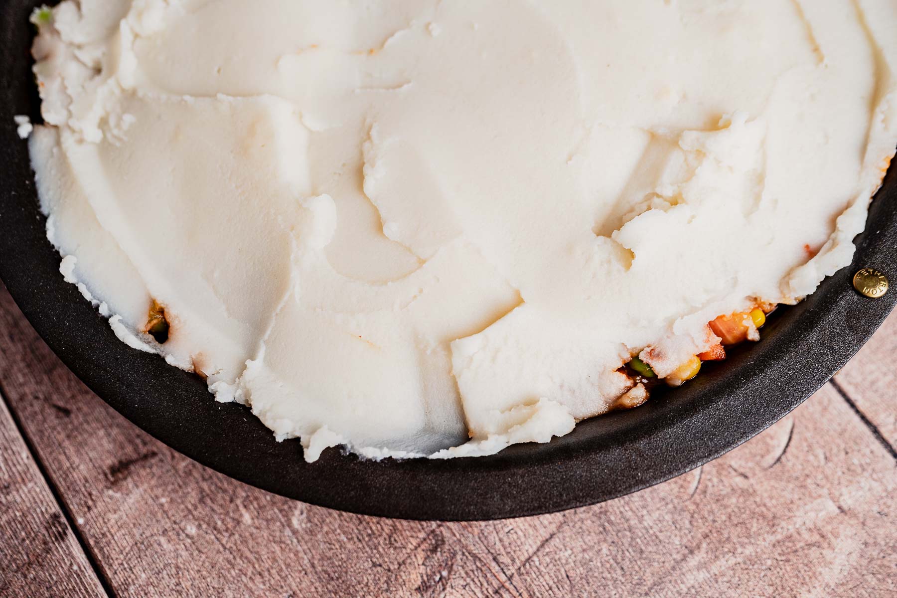 A close-up of an unbaked vegetarian shepherd's pie in a black round pan, topped with a smooth layer of mashed potatoes, showing some filling along the edge.