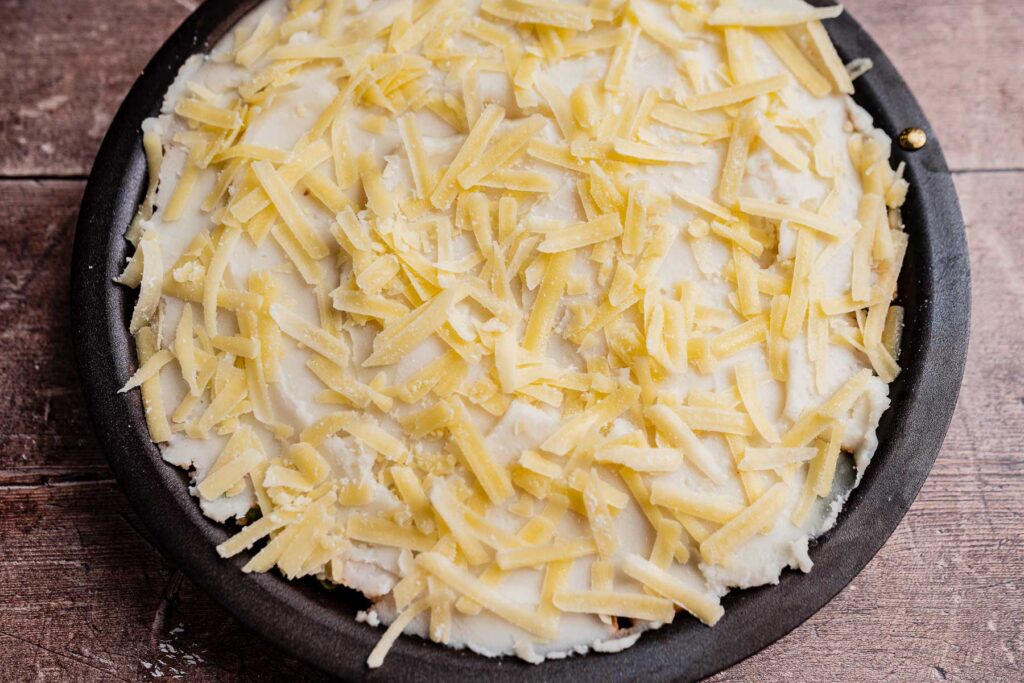 A round baking dish filled with an unbaked vegetarian shepherd's pie, topped with shredded cheese and a layer of mashed potatoes, on a wooden surface.