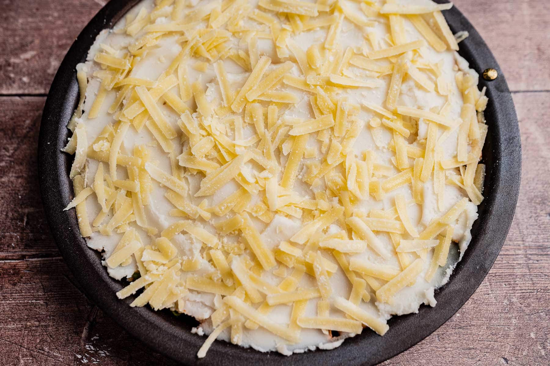 A round baking dish filled with an unbaked vegetarian shepherd's pie, topped with shredded cheese and a layer of mashed potatoes, on a wooden surface.